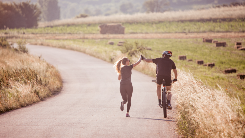 Runner and a cyclist high fiving on a road, surrounded by tall grass