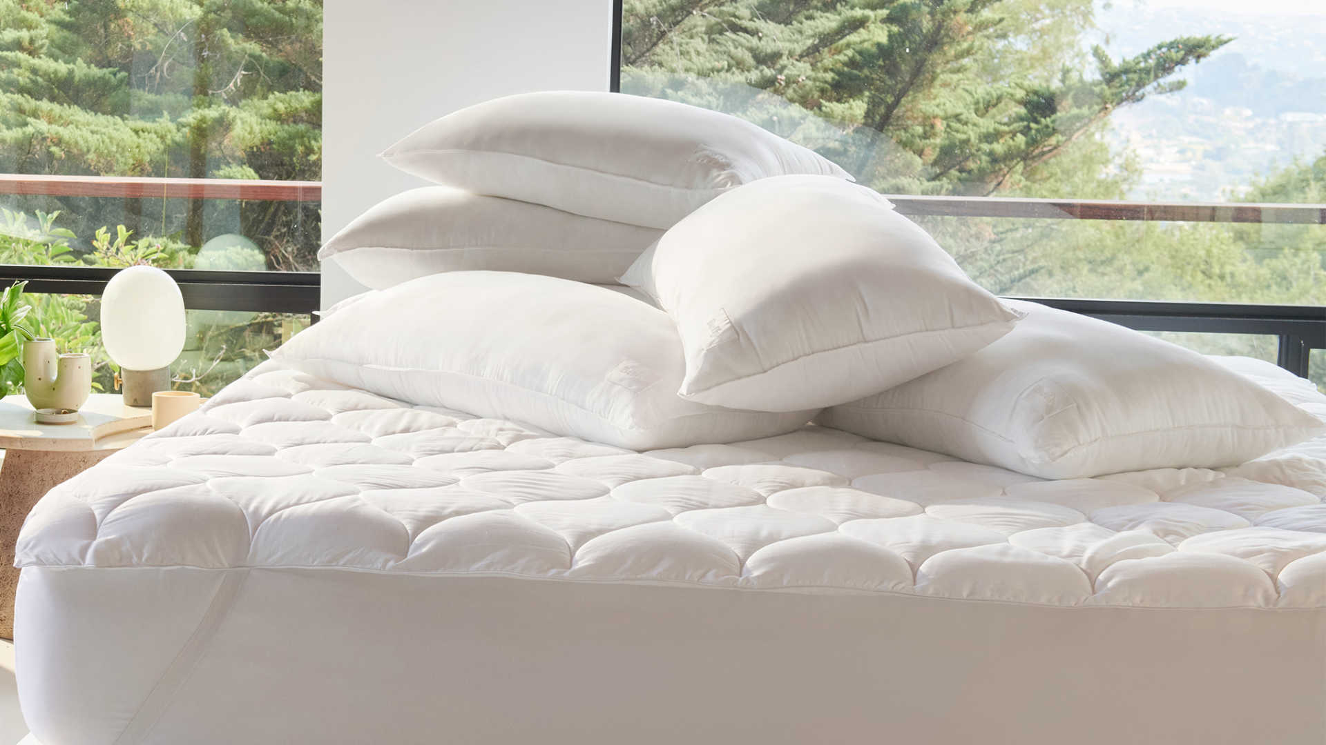 Unmade bed with pile of pillows and wall of windows with trees outside