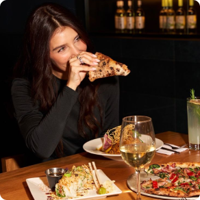 Young woman eats woodfired pizza in a dark restaurant at a table with a glass of wine