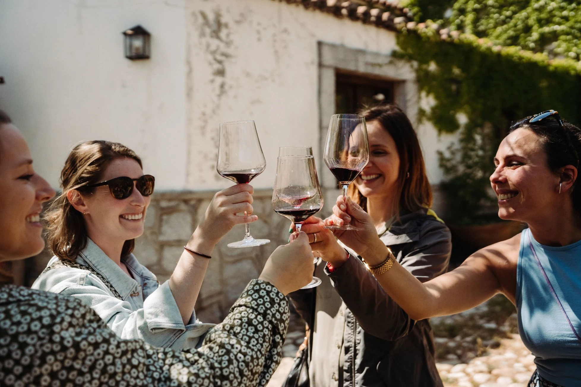 A group of four women outdoors raising glasses of red wine in a toast at a sunny vineyard or courtyard, smiling and enjoying a wine tasting experience.