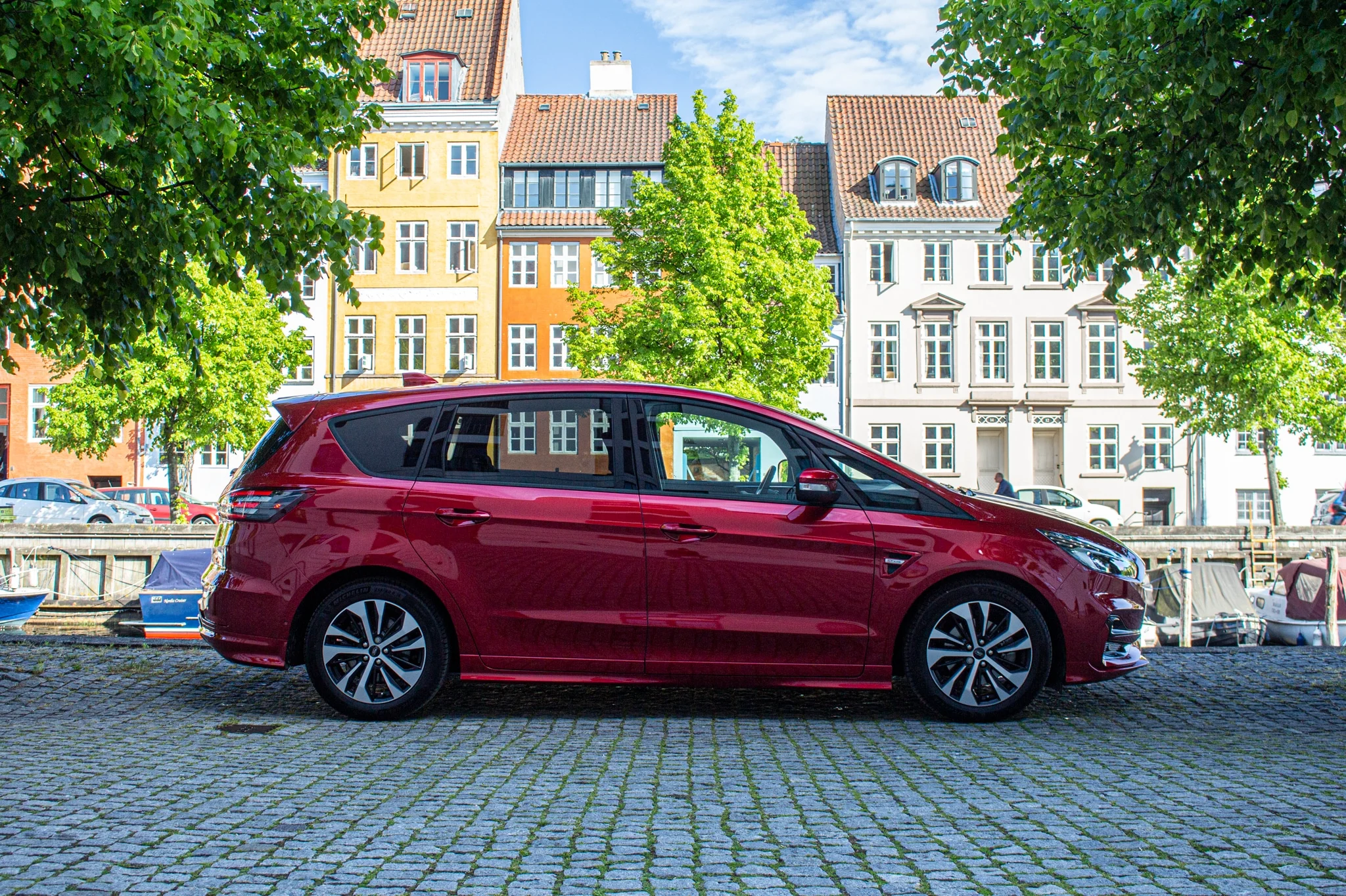 Röd kombi parkerad på kullersten bredvid å med fasader i bakgrunden