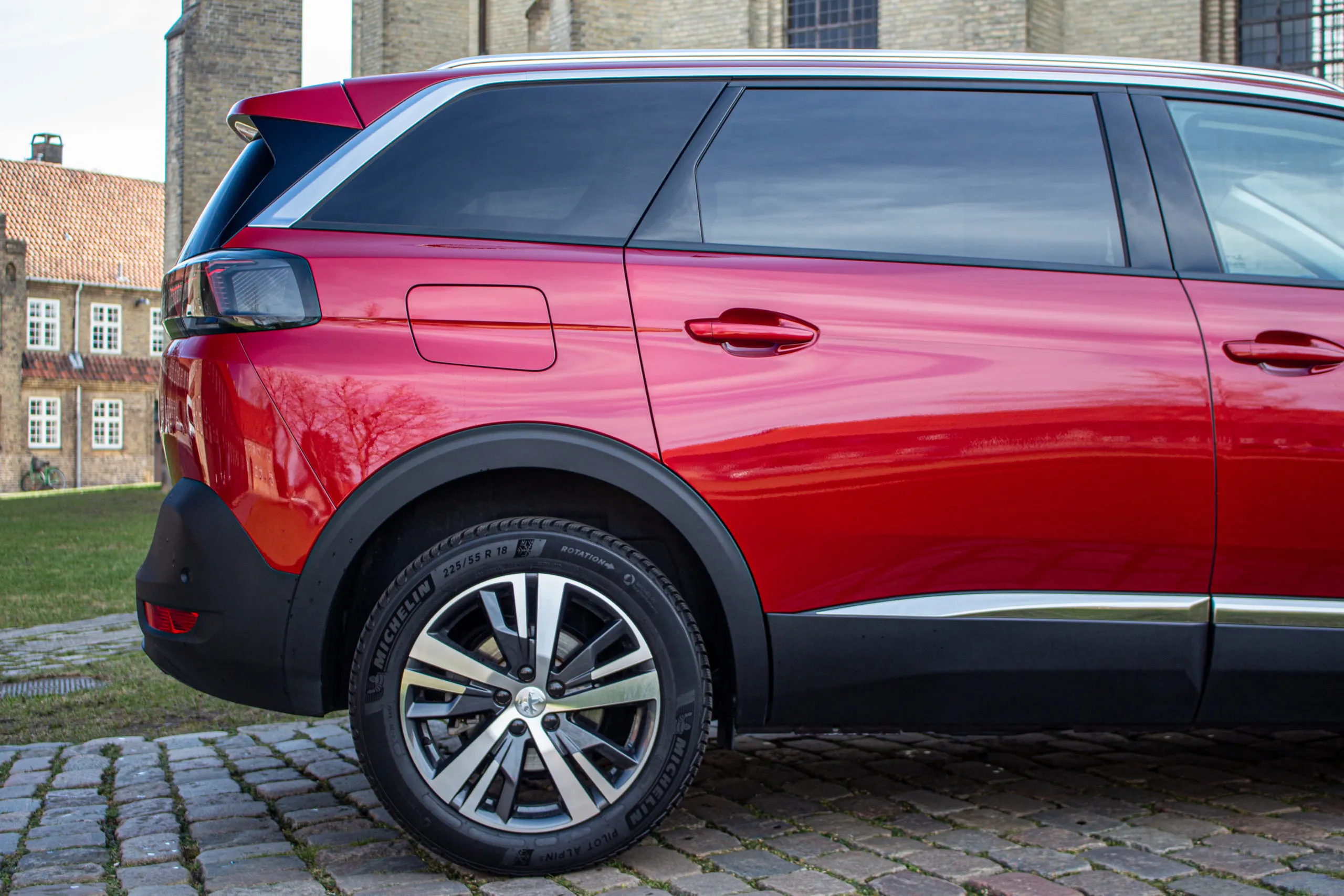 Baksidan av en röd Peugeot 5008 parkerad på en stenlagd yta framför en historisk byggnad.