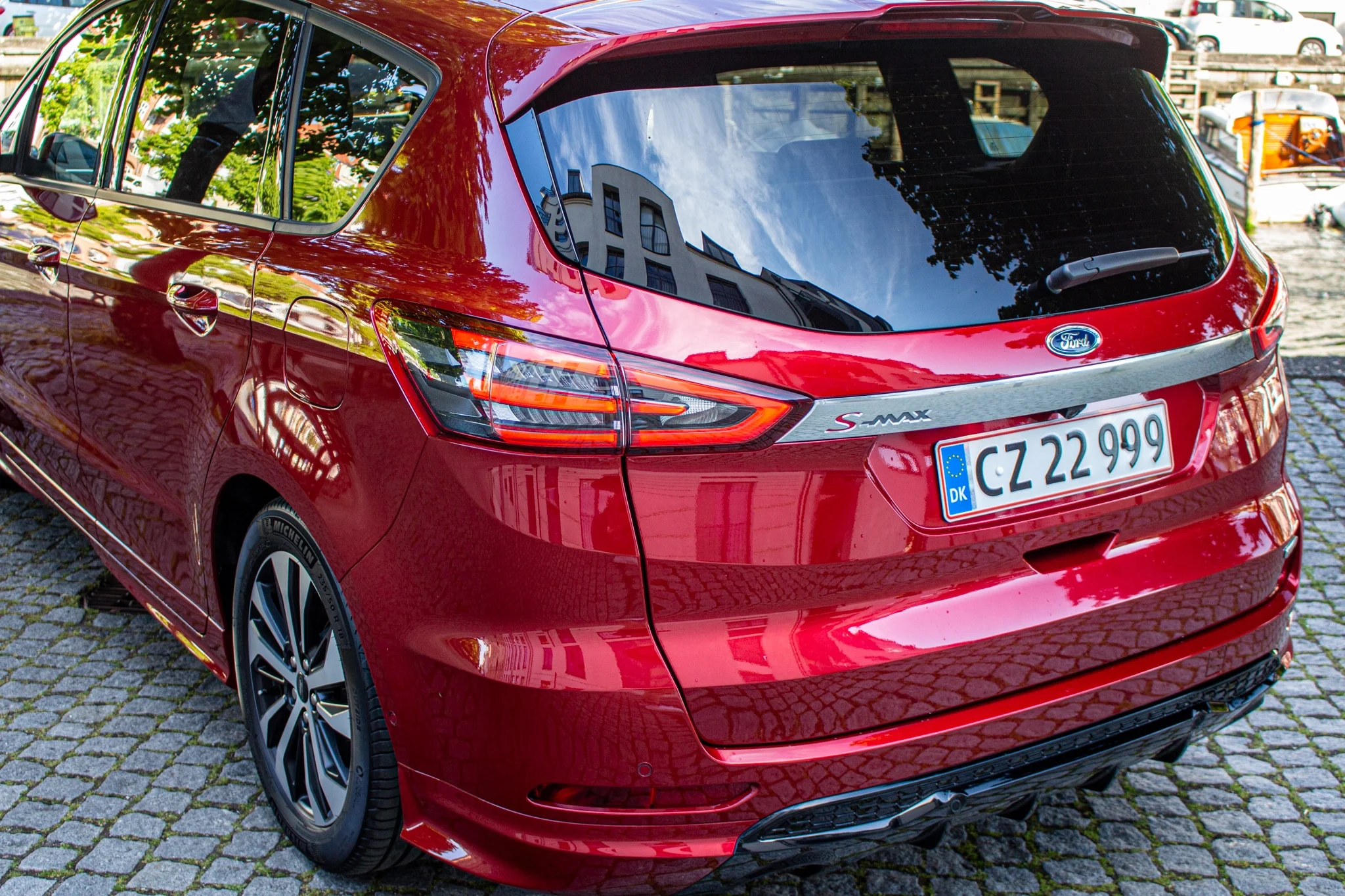 Baksidan av en röd Ford S-Max Hybrid parkerad på en stenlagd yta med reflektioner av omgivande byggnader i fönstren.