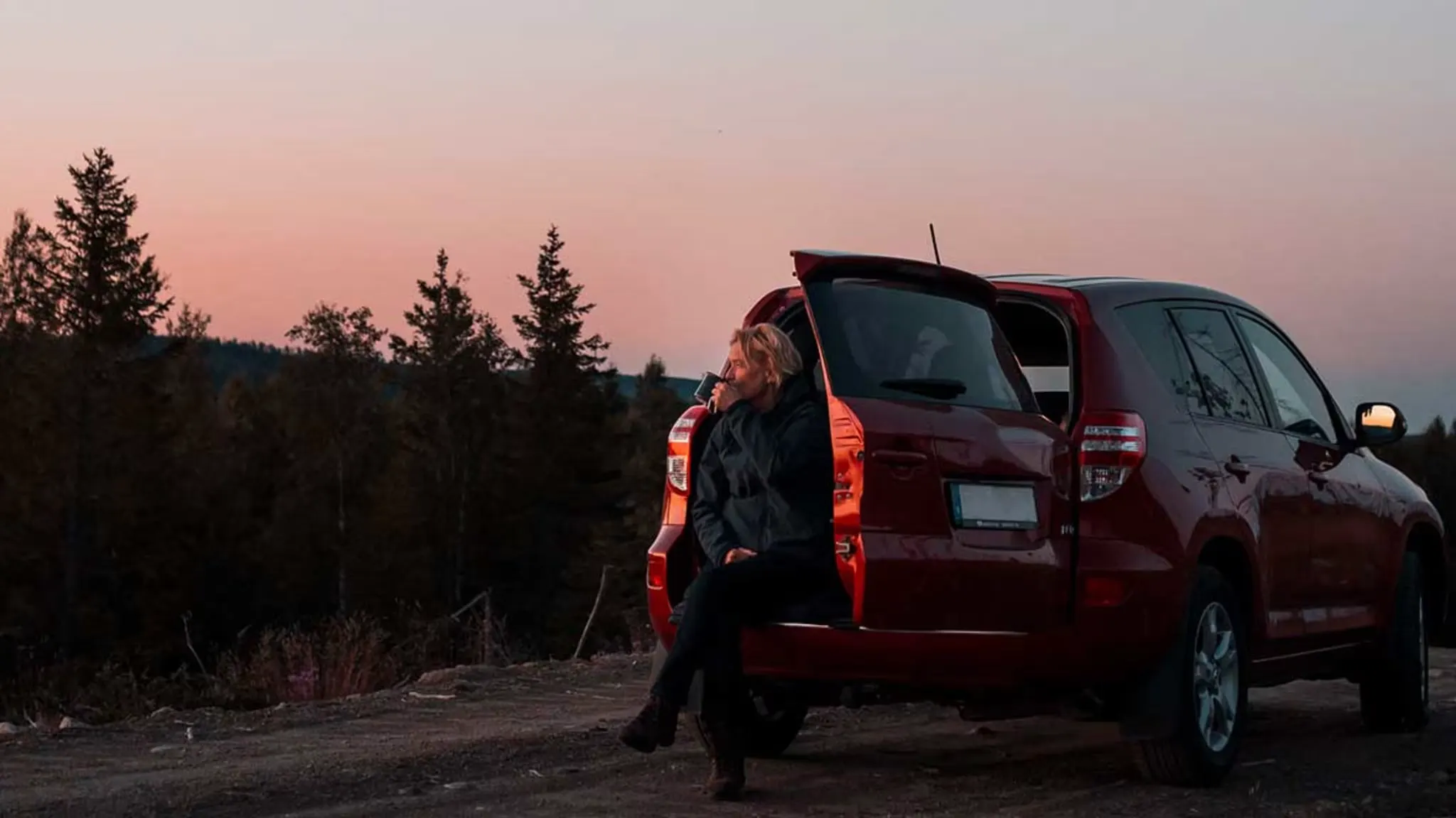 Kvinna sitter i på bakluckan av rödbil vid skymning 