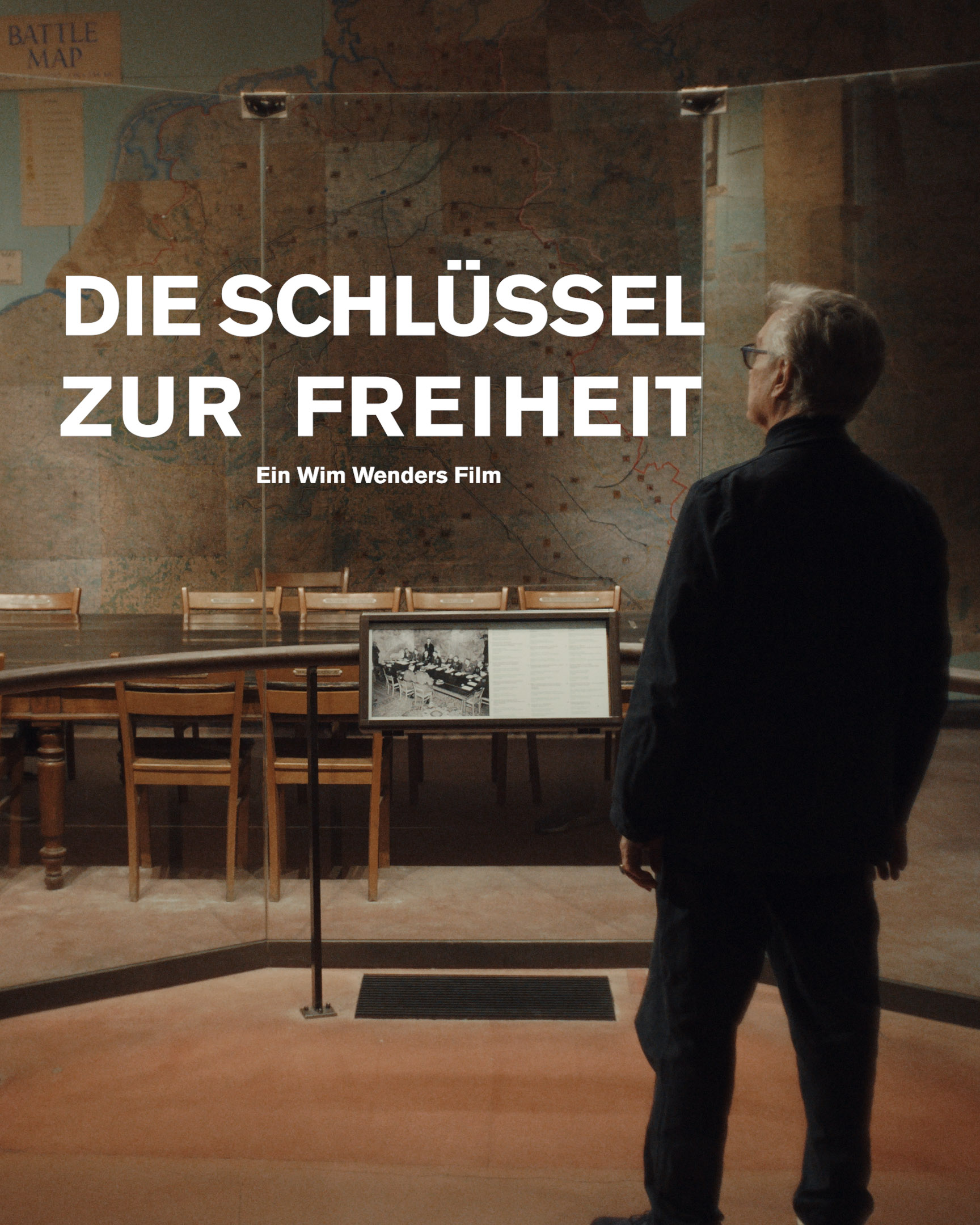 Ein Mann steht in einem Museum und blickt auf einen Konferenzraum mit langem Tisch und Stühlen. An der Wand dahinter sind große Landkarten aus Kriegszeiten zu sehen. Im Vordergrund steht: „Die Schlüssel zur Freiheit. Ein Wim Wenders Film.“