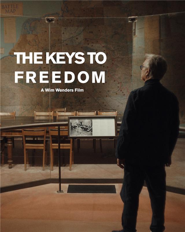 A man stands in a museum and looks at a conference room with a long table and chairs. Large maps from wartime are displayed on the wall behind him. In the foreground is written: “The Keys to Freedom. A Wim Wenders film.”