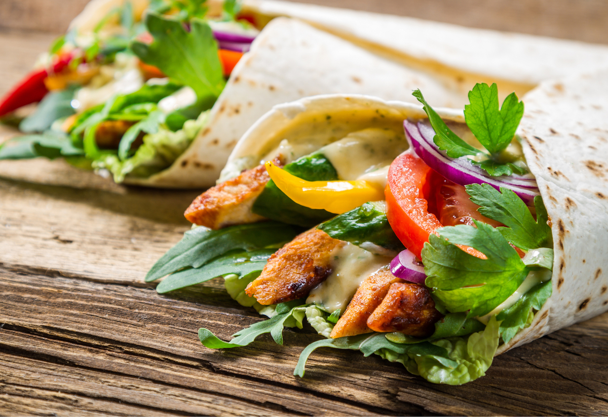 A fresh wrap bursting with lettuce, yellow and red peppers, chicken and sauces, sitting on a wooden backdrop