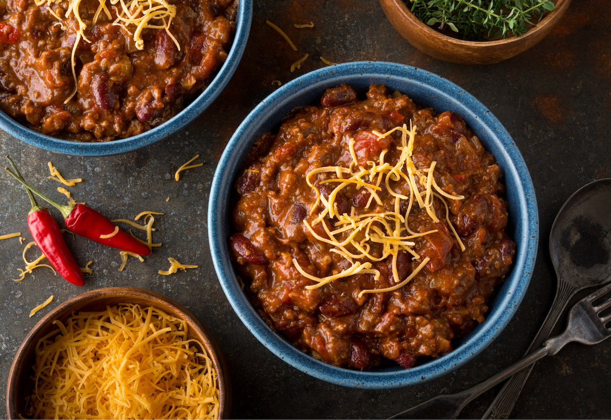 A bowl of piping-hot chili with grated cheese on top and two hot peppers nearby