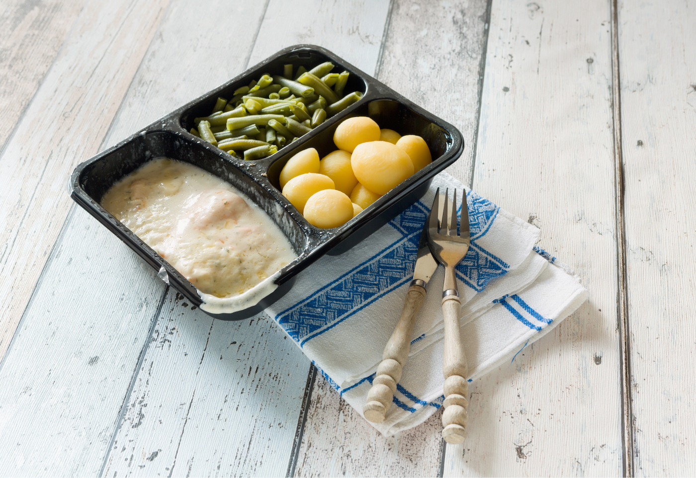 A frozen meal of beans, potatoes and meat sit near utensils and napkins on a wooden backdrop