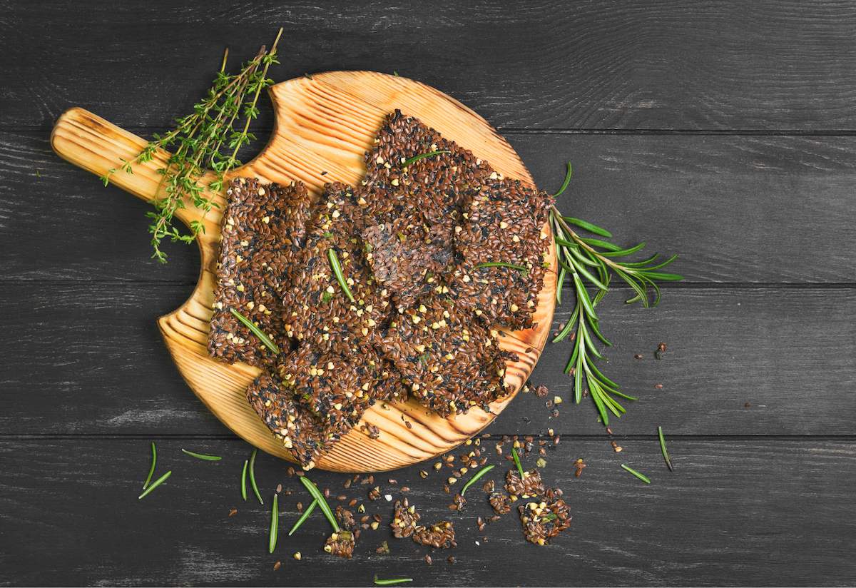 A wooden platter piled with deep-brown gluten-free crackers and crumbled herbs