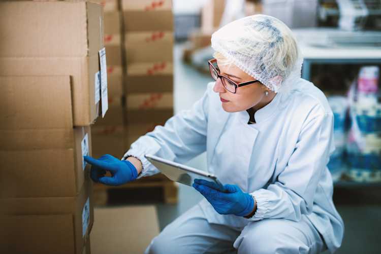A woman in glasses and a hairnet holds a clipboard and inspects cardboard boxes