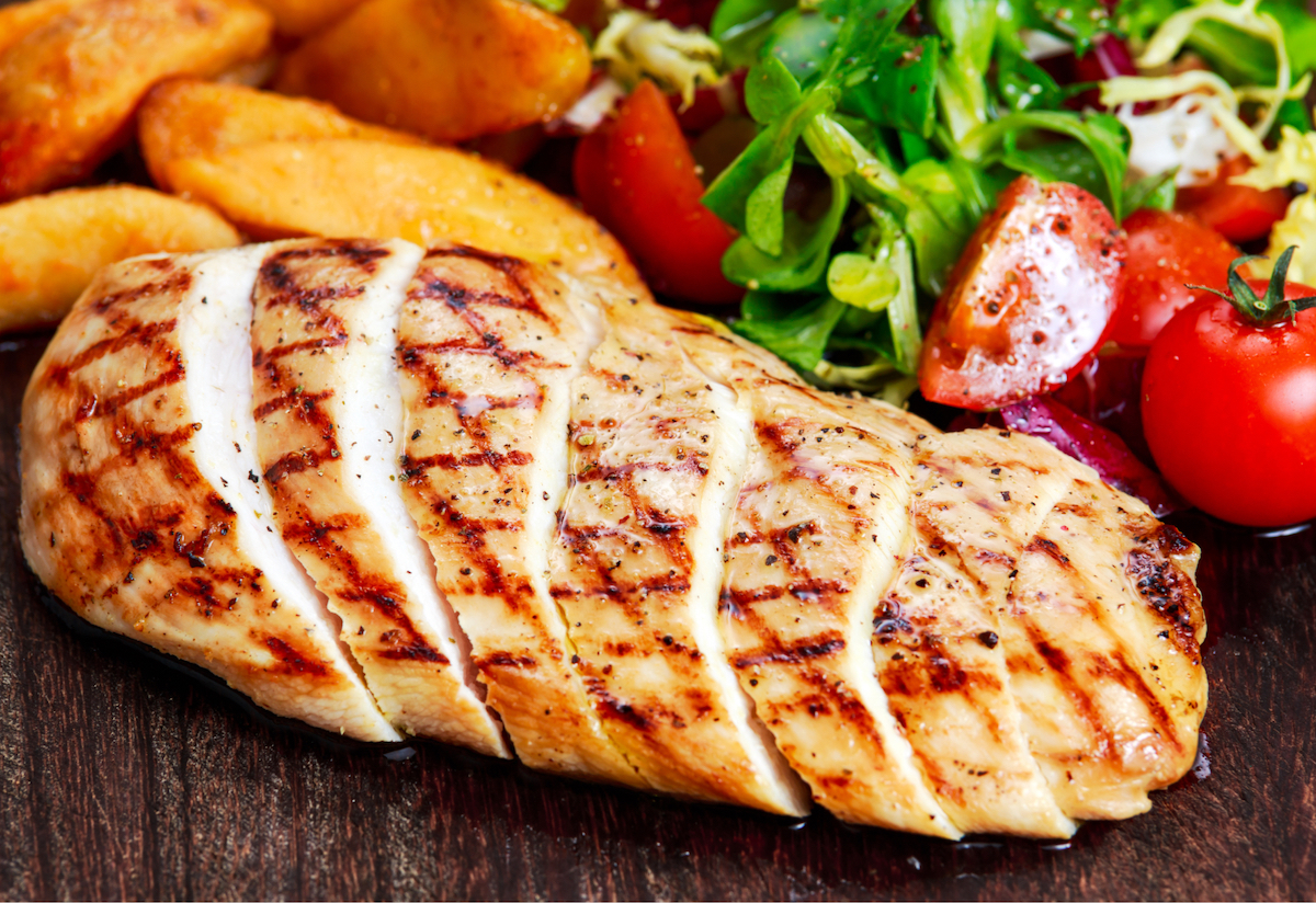 A sliced cooked chicken sits on a wooden table with tomatoes, lettuce and cut vegetables