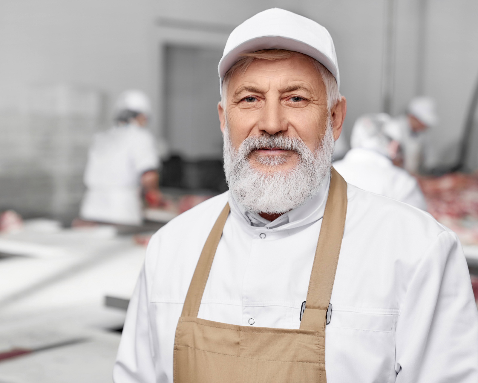 A bearded man with a white hat and beige apron smiles kindly at the camera