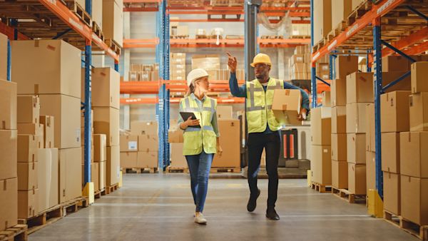 Two workers in yellow jackets and jeans walk down a hallway lined with large cardboard boxes.