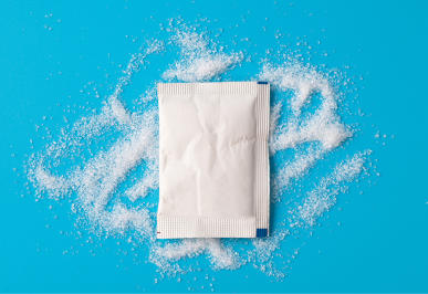 A white, unopened packet of sugar sits on sugar sprinkled over a blue backdrop