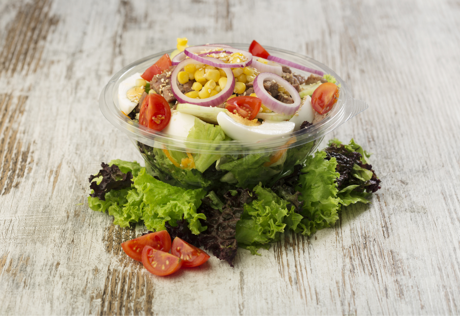 A cobb salad in a clear plastic bowl, on a wooden backdrop
