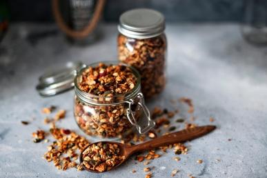 Fresh granola in a glass jar, pouring out into a wooden spoon on a grey backdrop