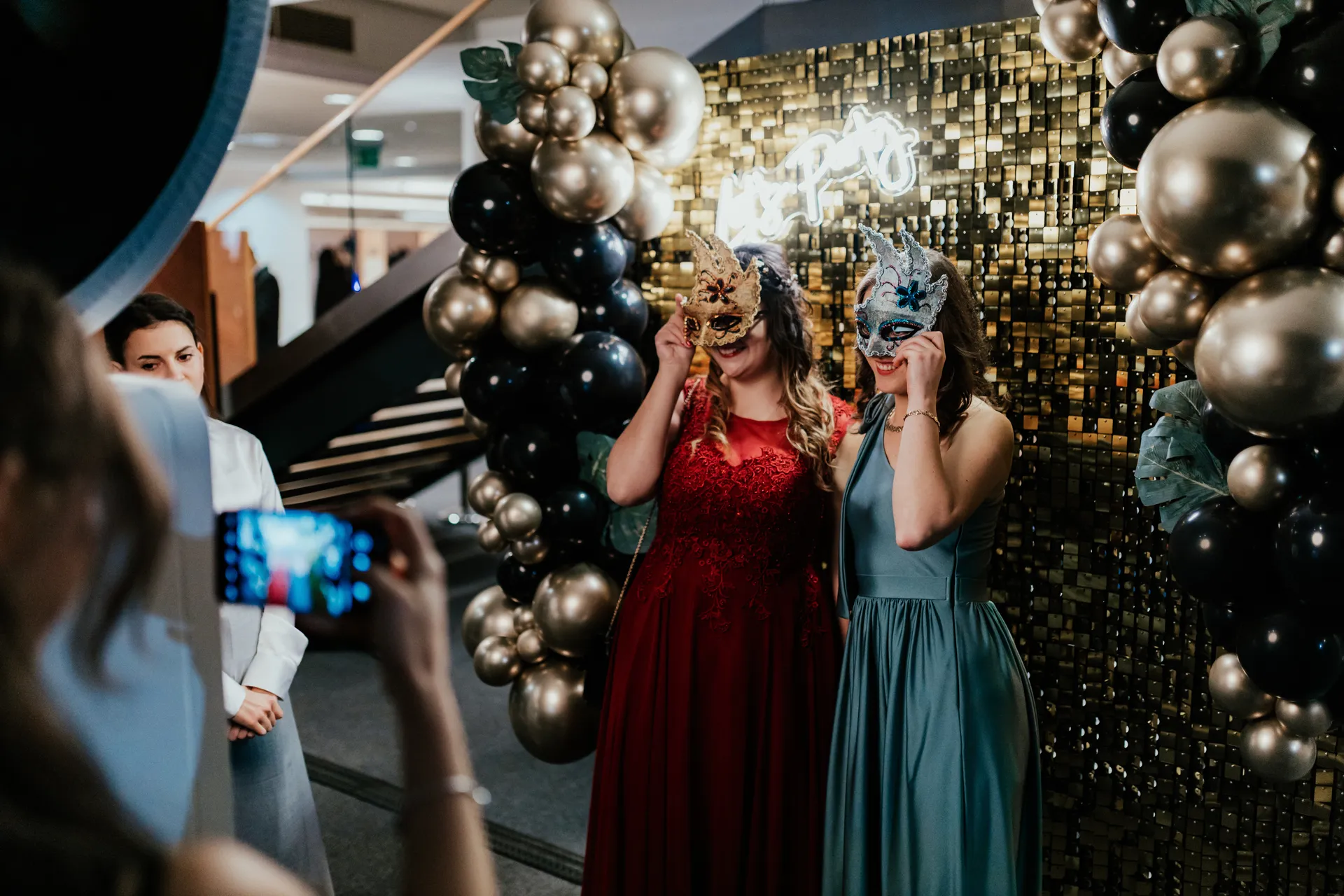 Estélyis hölgyek maszkban pózolnak a prémium lufis fotófal és a szelfigép előtt egy elegáns bálon.