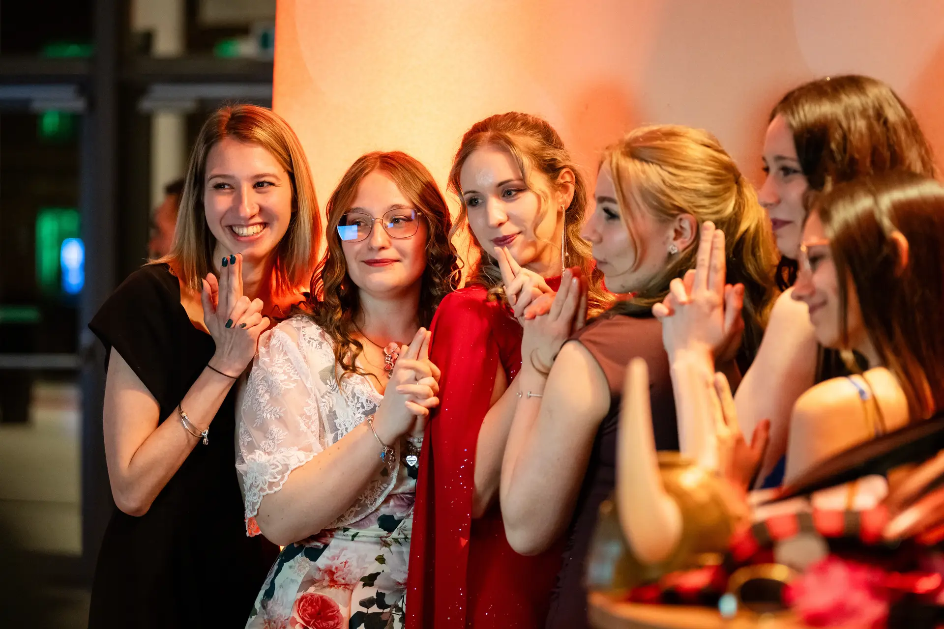 Elegáns, mosolygó lányok pózolnak az esküvői fotósarok előtt, vicces kellékekkel az előtérben.