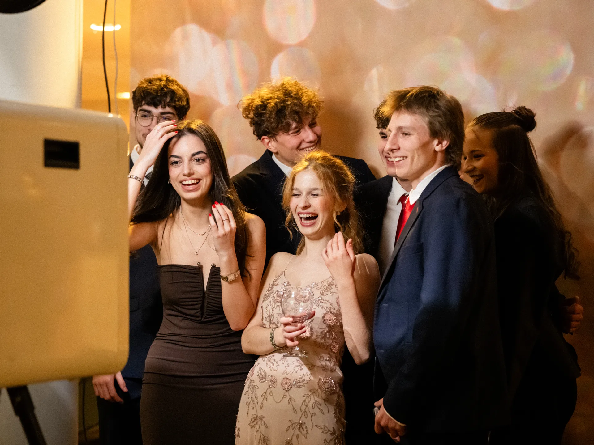 Önfeledten nevető, elegáns fiatalok pózolnak a képnyomtatós automata előtt egy nívós rendezvényen.