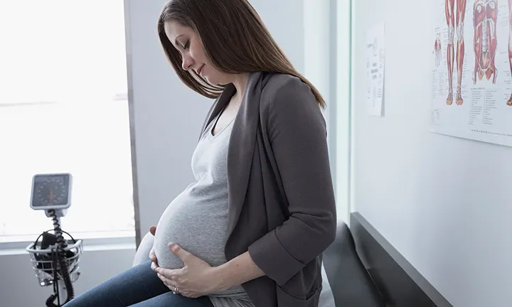 Pregnant woman sitting on a doctor's exam table, holding her belly, possibly preparing for an epidural during labor.