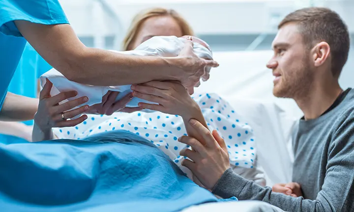 A mother in a hospital bed receives her newborn from a healthcare professional, with her partner beside her.