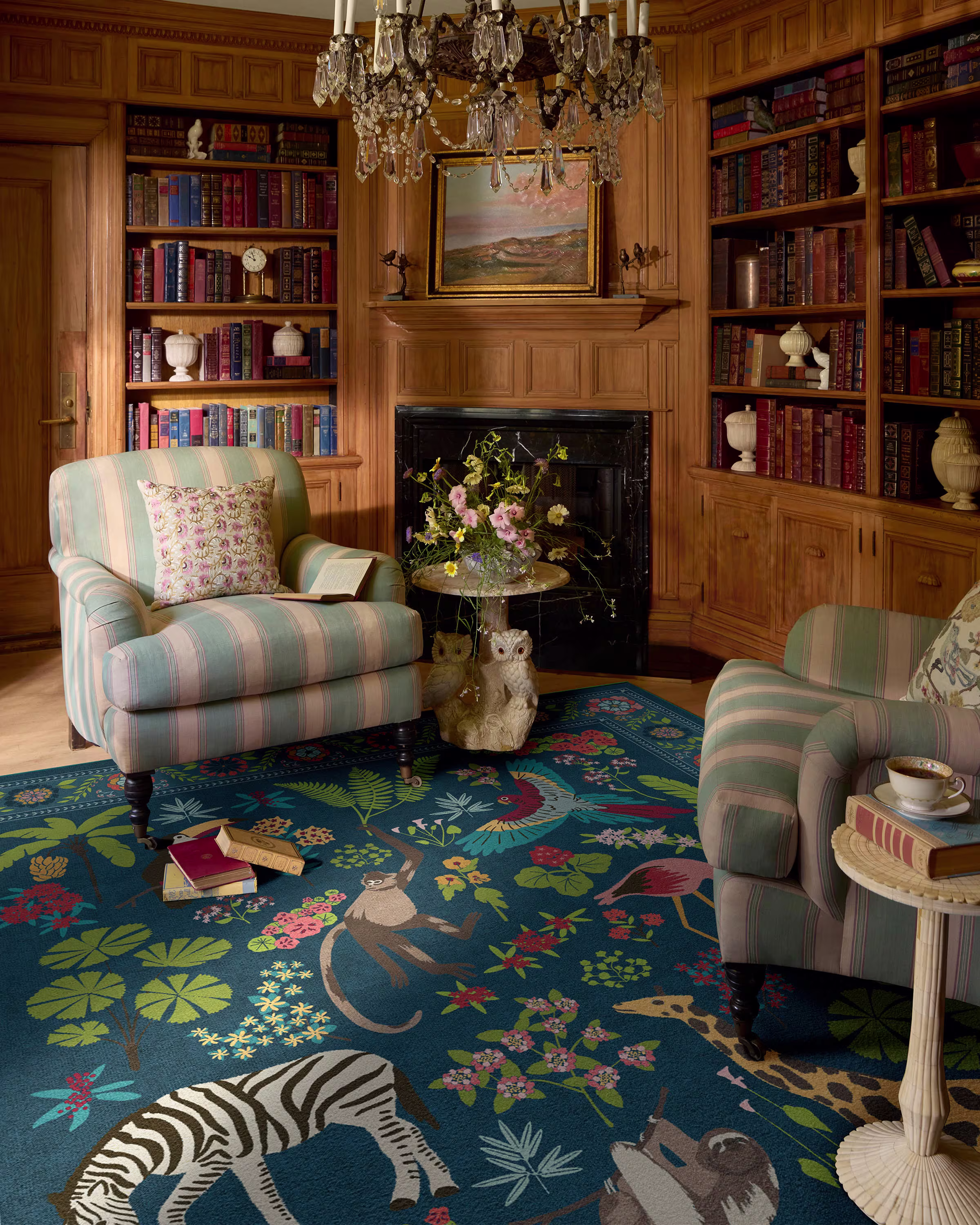 A vibrant royal blue Iris Apfel Jingle Jungle flatwoven area rug featuring illustrations of zebras, monkeys, and tropical flora, placed in a traditional wood-paneled home library with striped armchairs and a fireplace.