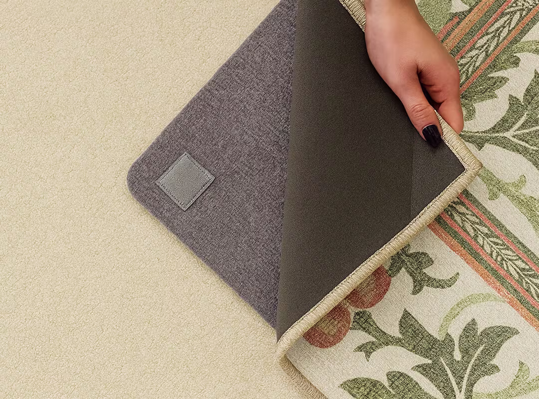 A person lifts the corner of Ruggable's Strawberry Thief Sage & Rose Rug to show the rug pad underneath, demonstrating how to use a rug pad when placing the rug on a carpet.