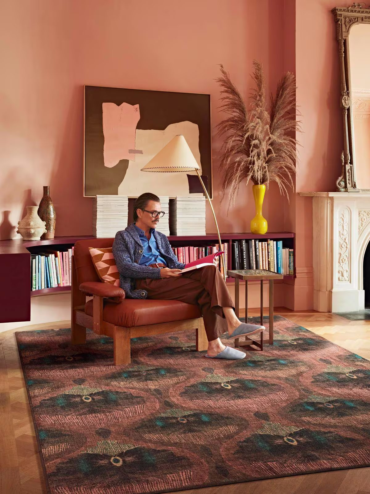 Image of interior designer Matthew Williamson sitting in a reading nook with a leather chair, bookcase, and the Matthew Williamson Pluma Rosewood Rug.