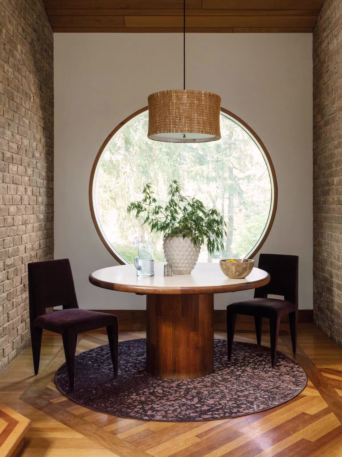 Image of a small dining nook in a room with patterned wood flooring, an exposed brick wall, a large circular window, round bistro table, and the round Ruggable x Architectural Digest Terran Burgundy Rug. 