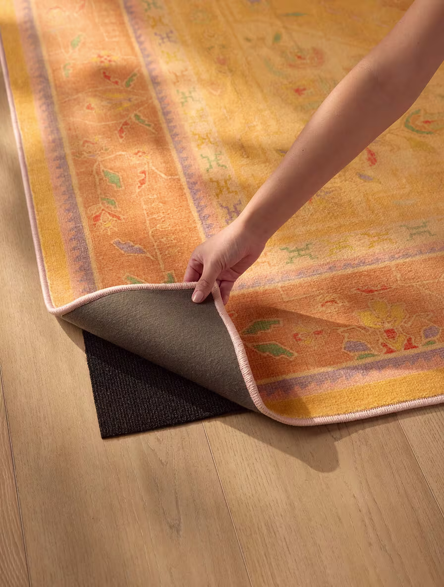 Hand lifting the corner of Ruggable's Colourful Loom Mango Rug to show the rug pad underneath, which helps prevent dents from heavy furniture.