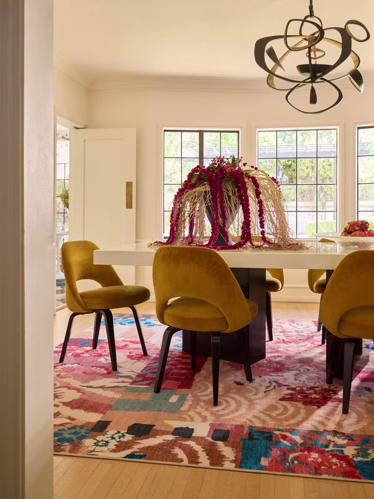 Image of sunny dining room with a white table, overflowing flower vase, yellow velvet chairs and the Ruggable x Iris Apfel Patchwork Peach Multicolor Rug.