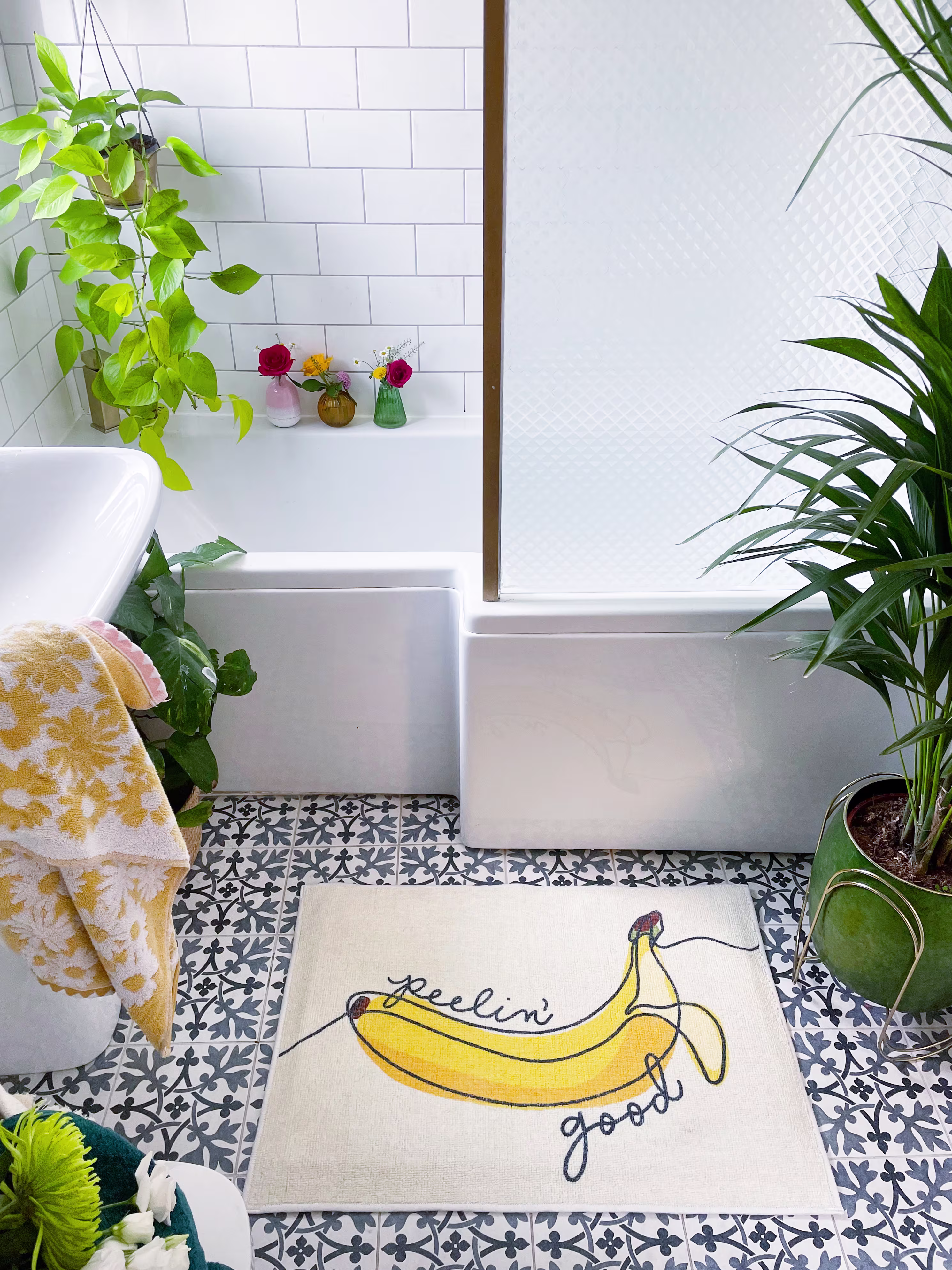 Ruggable Peelin' Good Novelty bath mat styled in an eclectic small bathroom featuring black and white patterned tile floors, white subway tile walls, a bathtub with a glass door, and vibrant green potted plants.