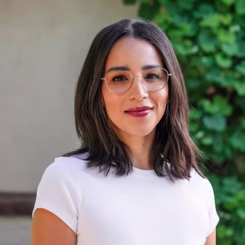 Person with shoulder-length dark hair wearing glasses and a white t-shirt, standing in front of green foliage.