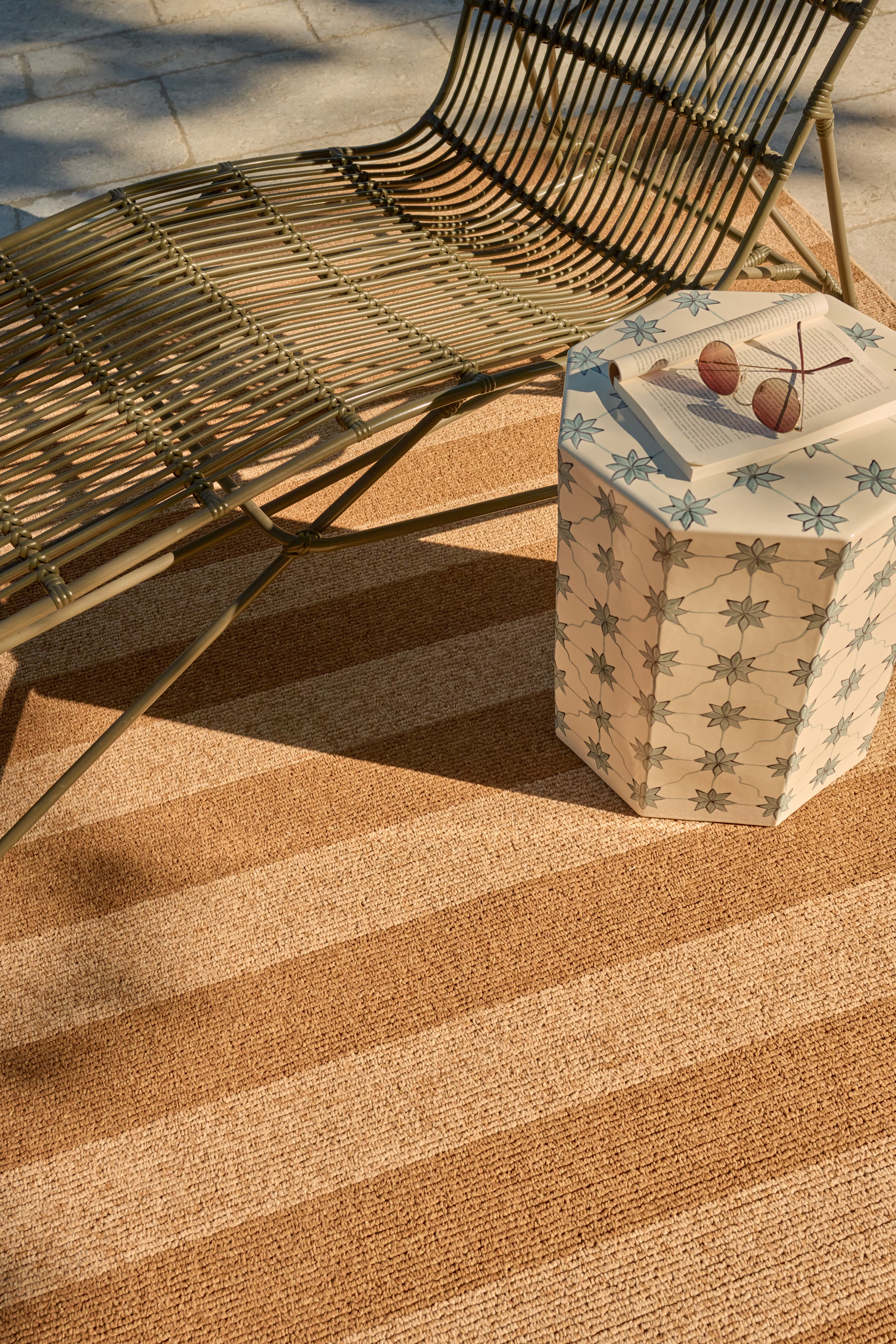 A high-angle view of the Ruggable Nolan Bold Stripe Performance Weave rug in natural and dark natural tones, styled poolside under a modern wire outdoor lounge chair next to a geometric mosaic side table.