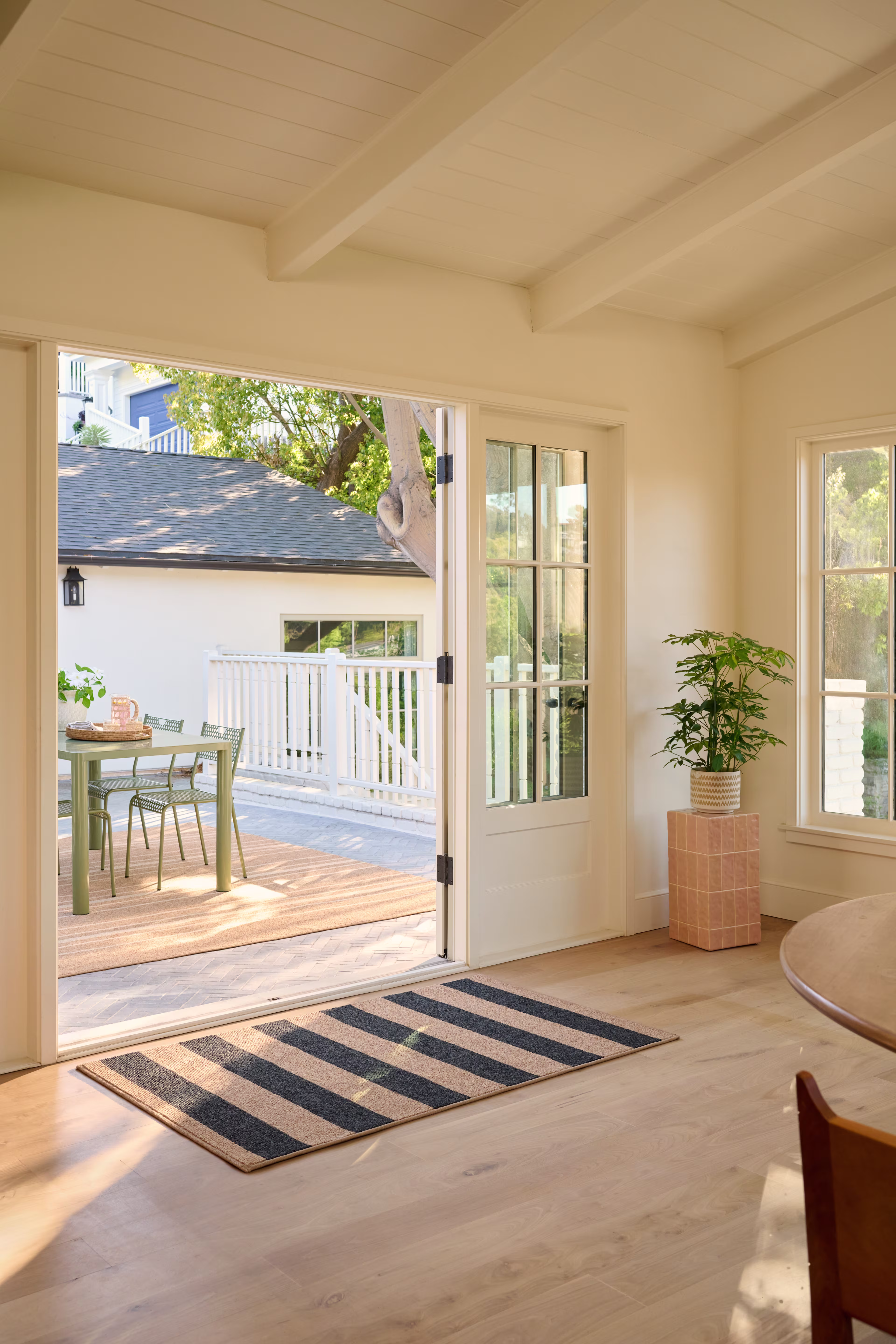A high-angle shot looking from a bright indoor room with light hardwood floors out through open French doors onto a sunny wood backyard patio. A small black and tan striped Ruggable Nolan Bold Stripe Performance Weave rug is placed neatly inside the doorway.