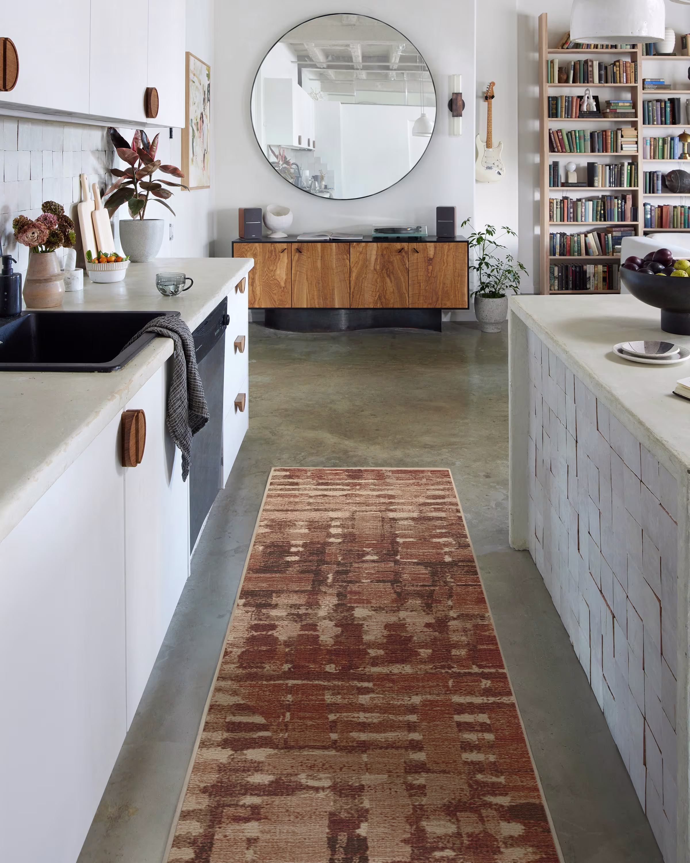 A long kitchen or hallway runner featuring Ruggable's Architectural Digest collection. The rust and copper-toned geometric pattern stretches down a polished concrete floor, beautifully contrasting with modern white kitchen cabinetry, black fixtures, and a large round vintage mirror hanging on the wall.