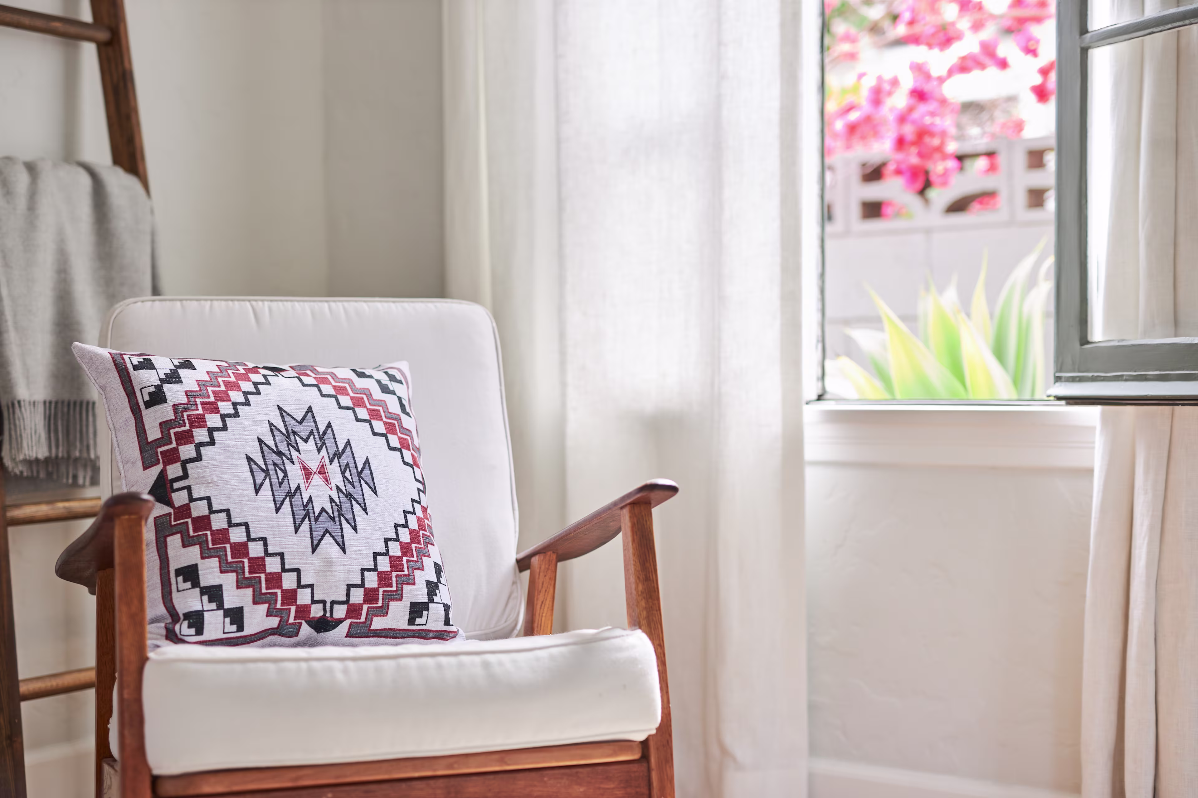 Wooden armchair with a cream cushion and a southwestern-patterned accent pillow, styled beside a sunlit window with soft curtains and greenery outside.