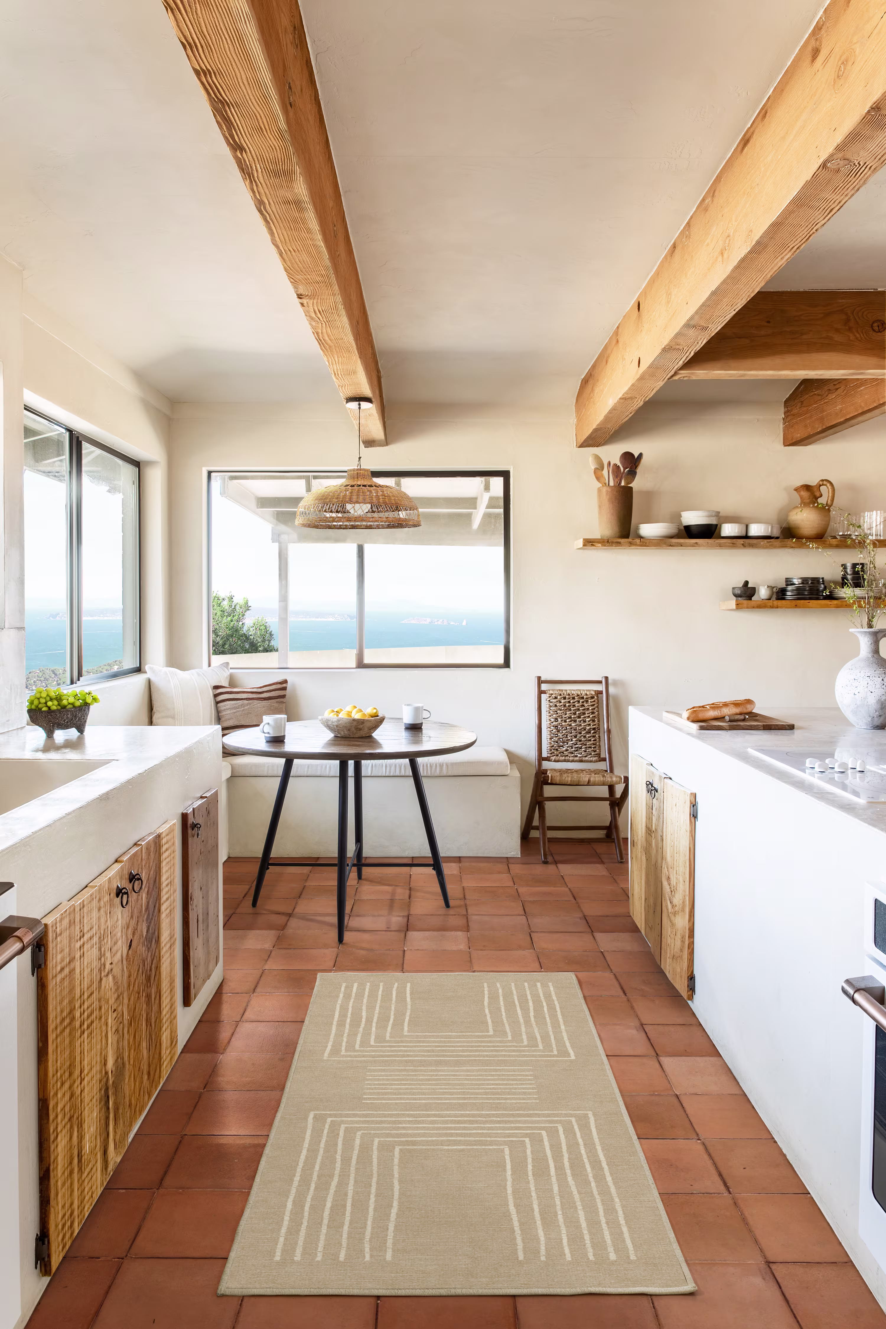 Cozy kitchen with terracotta tiles, wooden beams, and a large window. Features a round table, white cabinets, and rustic shelves, creating a warm, inviting atmosphere.