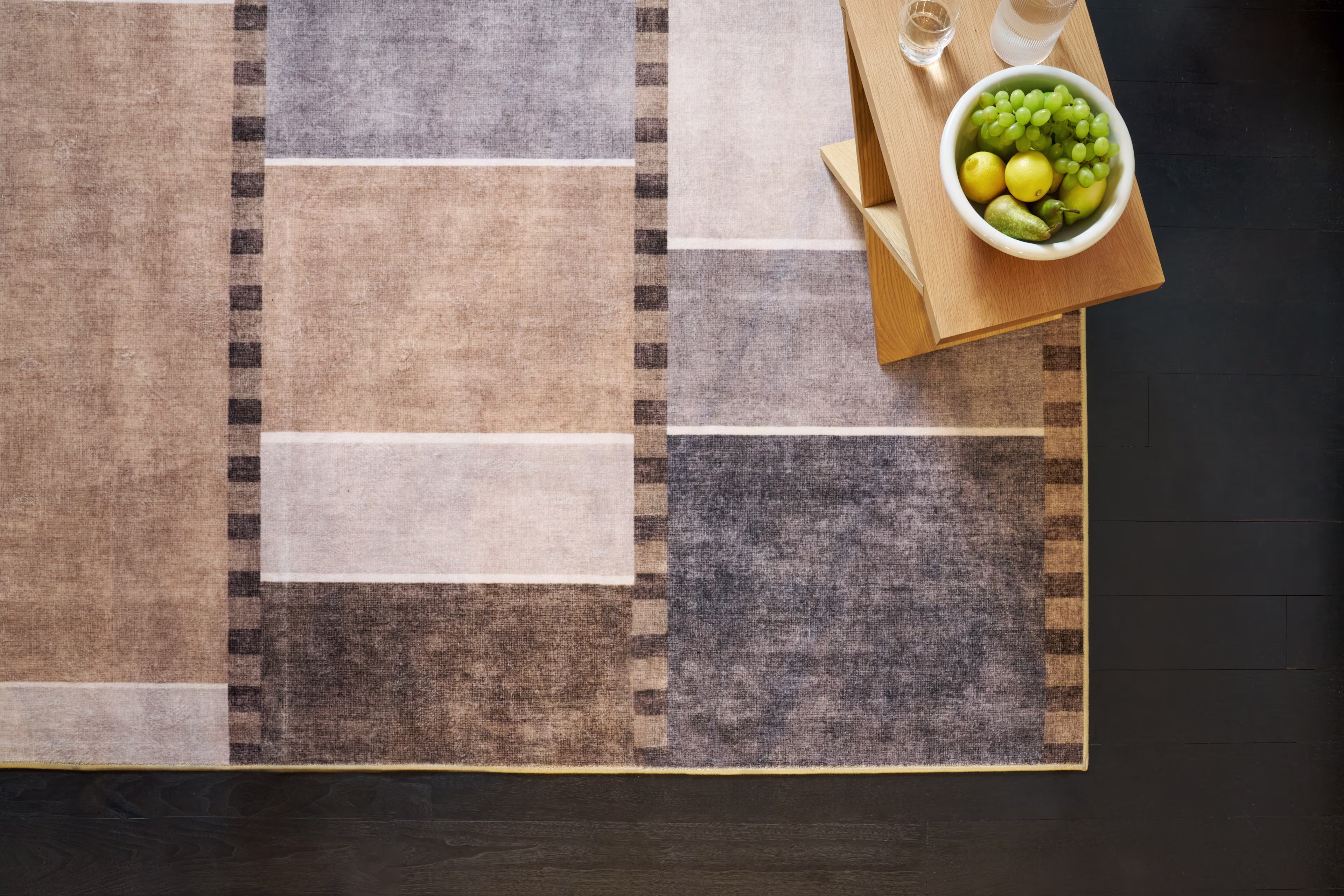 Top view of a modern geometric area rug in beige, gray, and brown tones with a wooden side table holding a bowl of green grapes and fruit on a dark wood floor.