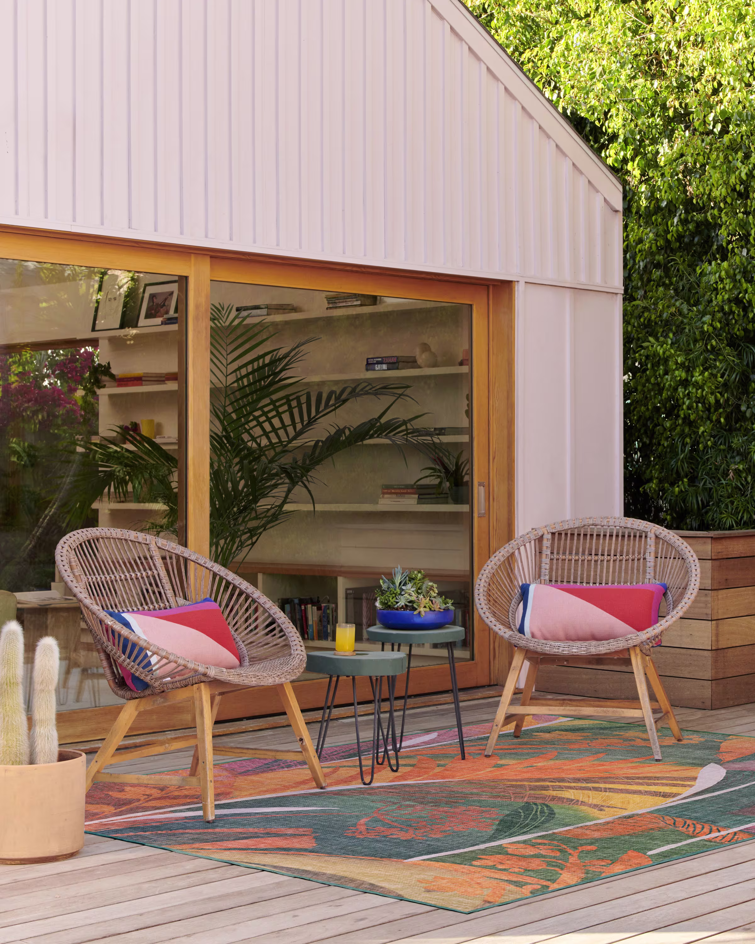 A vibrant, jungle-inspired Ruggable Palmera Multicolor outdoor rug styled on a wooden backyard deck. Two modern woven round accent chairs with colorful pillows flank a dark gray metal nesting side table holding drinks.