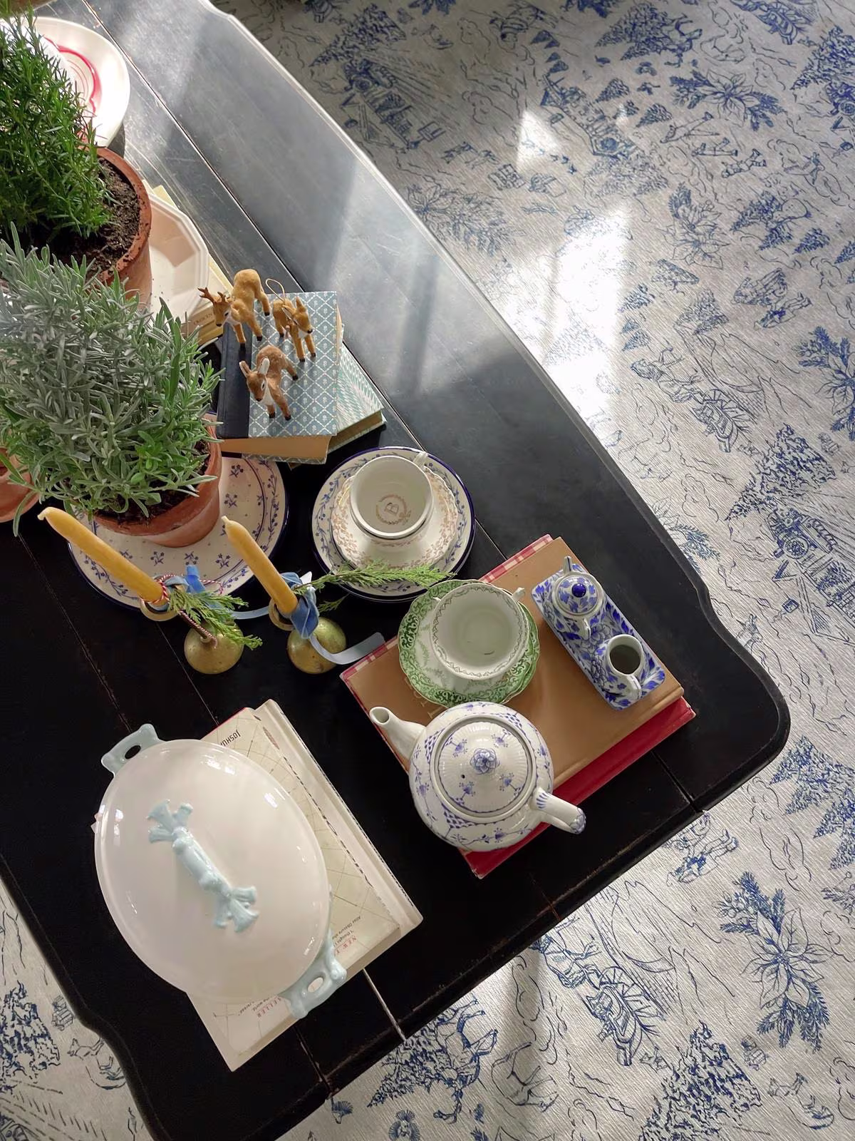 Image of a black wooden coffee table styled with small rosemary plants, a tea set, books, and Ruggable's Winter Toile Blue Rug underneath. 