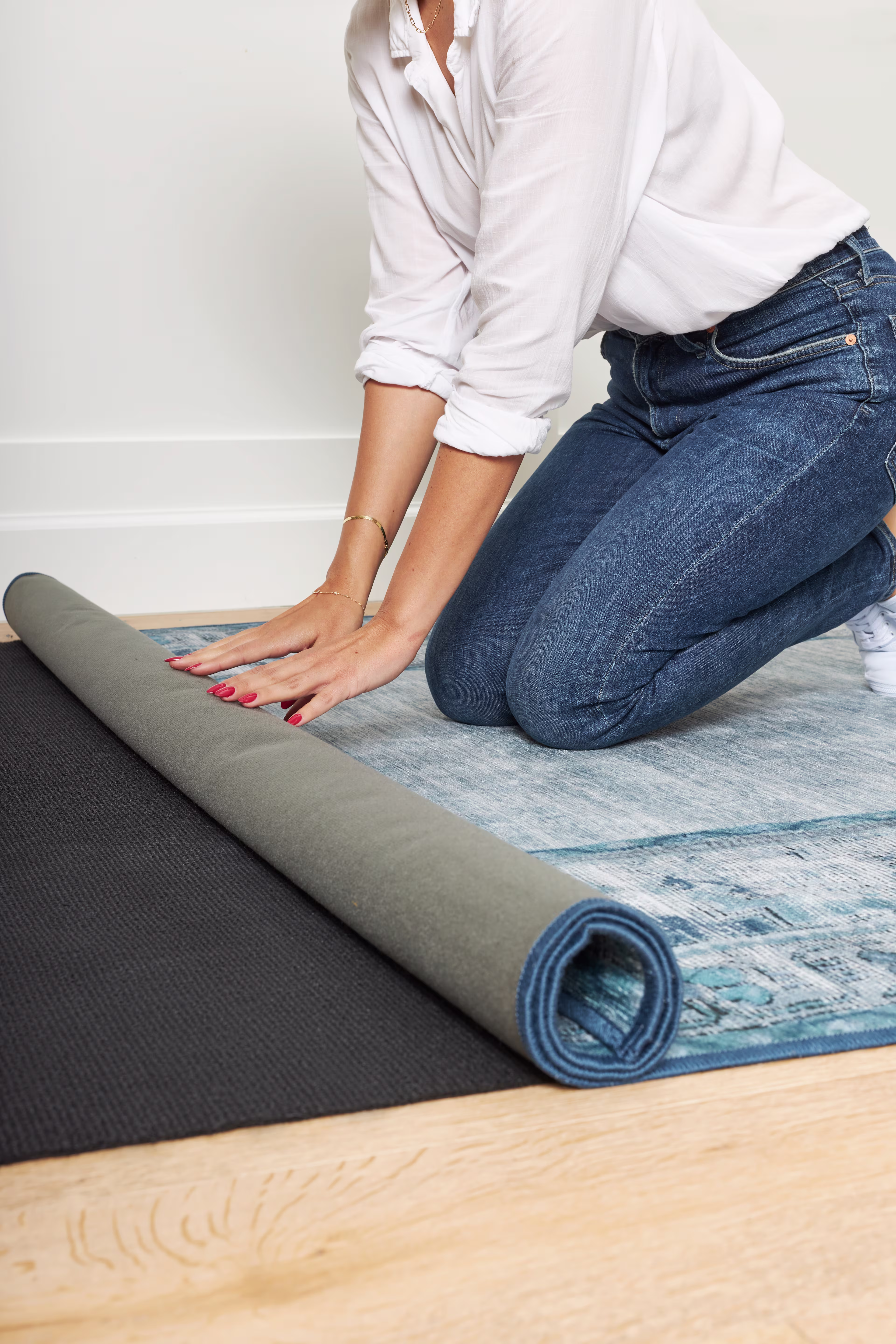 A woman on her knees unrolling a blue and green Vintage Daisy Bordered Ruggable rug over a black classic rug pad on light hardwood floors.