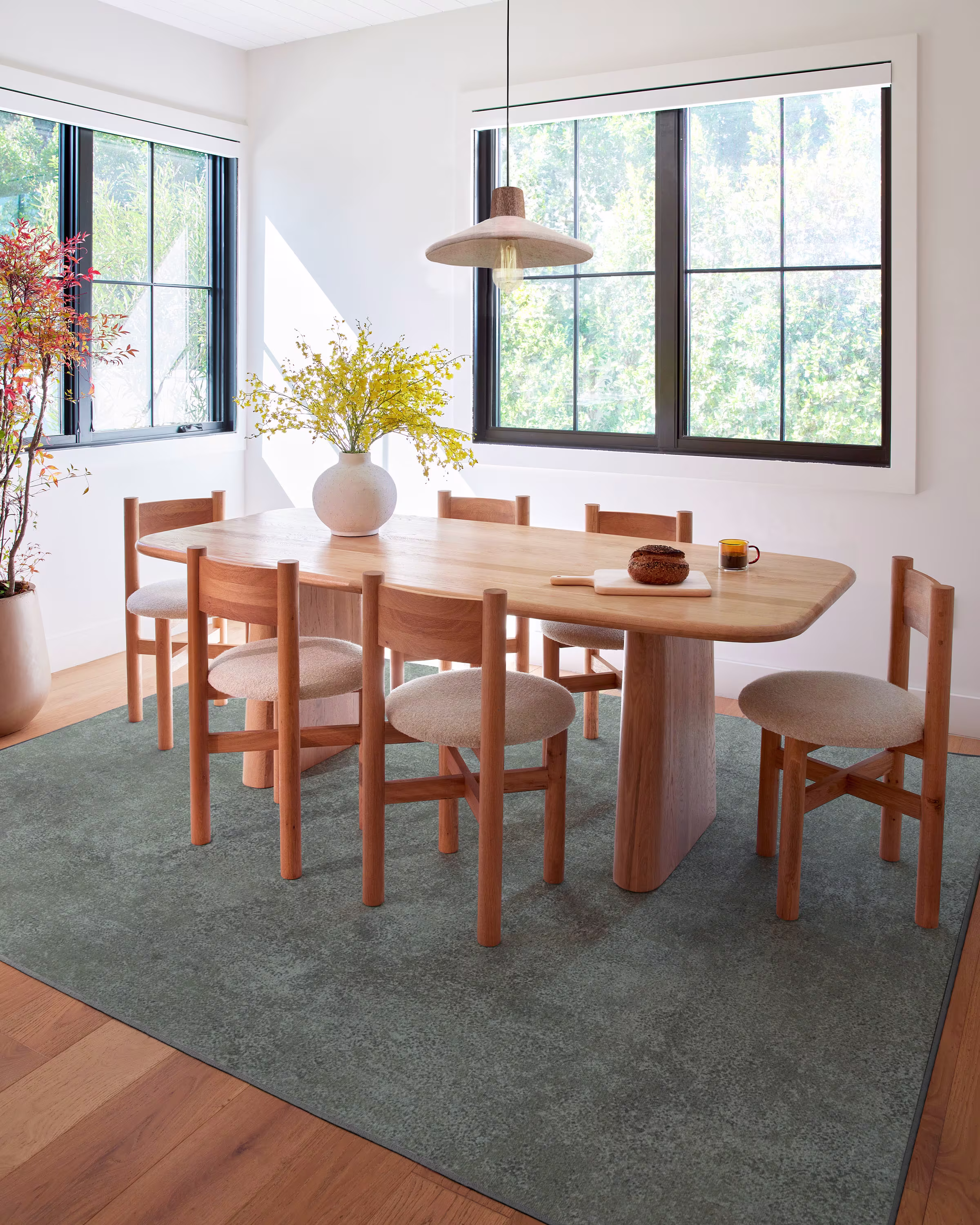 A modern dining room with a wooden table, six chairs, a vase of yellow flowers, a cup, a plate with bread, large windows, and a pendant light above the table.