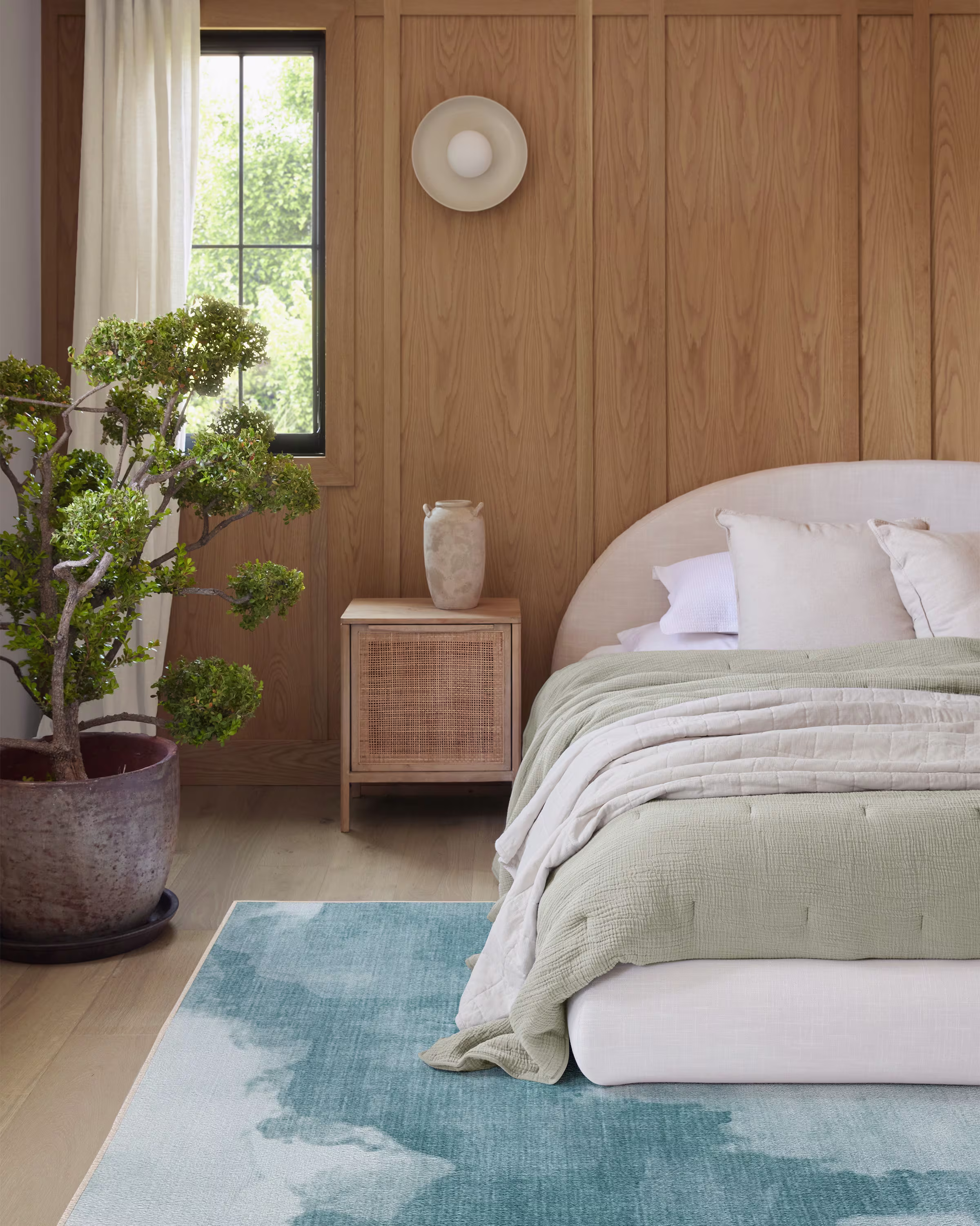 A modern bedroom with a light wood paneled wall, a bed with green and beige bedding, a potted plant, a small nightstand, and a blue area rug.