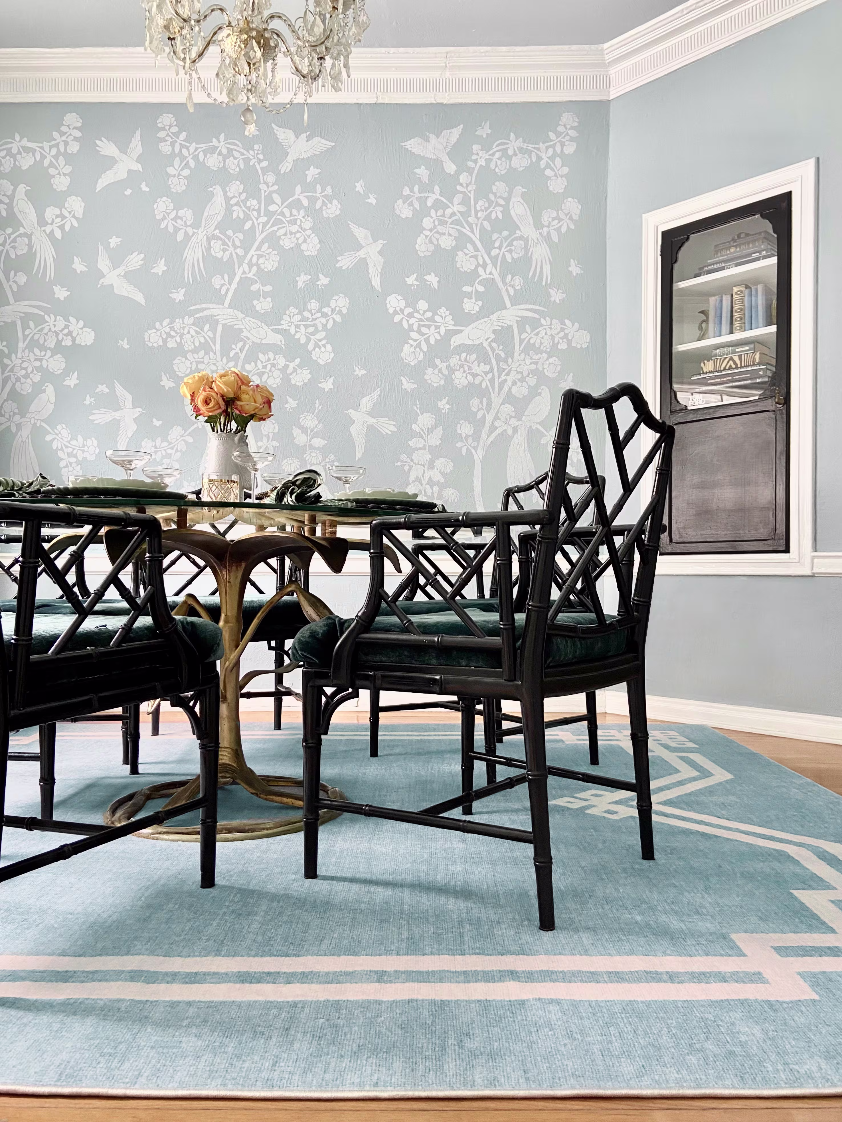 Elegant dining room with black lattice chairs, a glass-topped table, and a chandelier. Wallpaper with white floral and bird motifs adds charm.