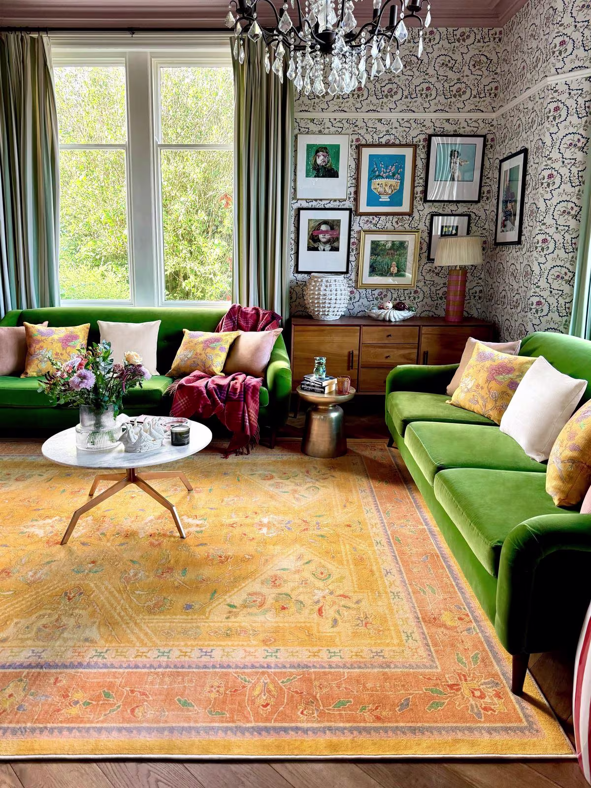 Image of luxurious living room with green velvet furniture, patterned wallpaper, crystal chandelier, and the Ruggable x Iris Apfel Colorful Loom Mango Rug.