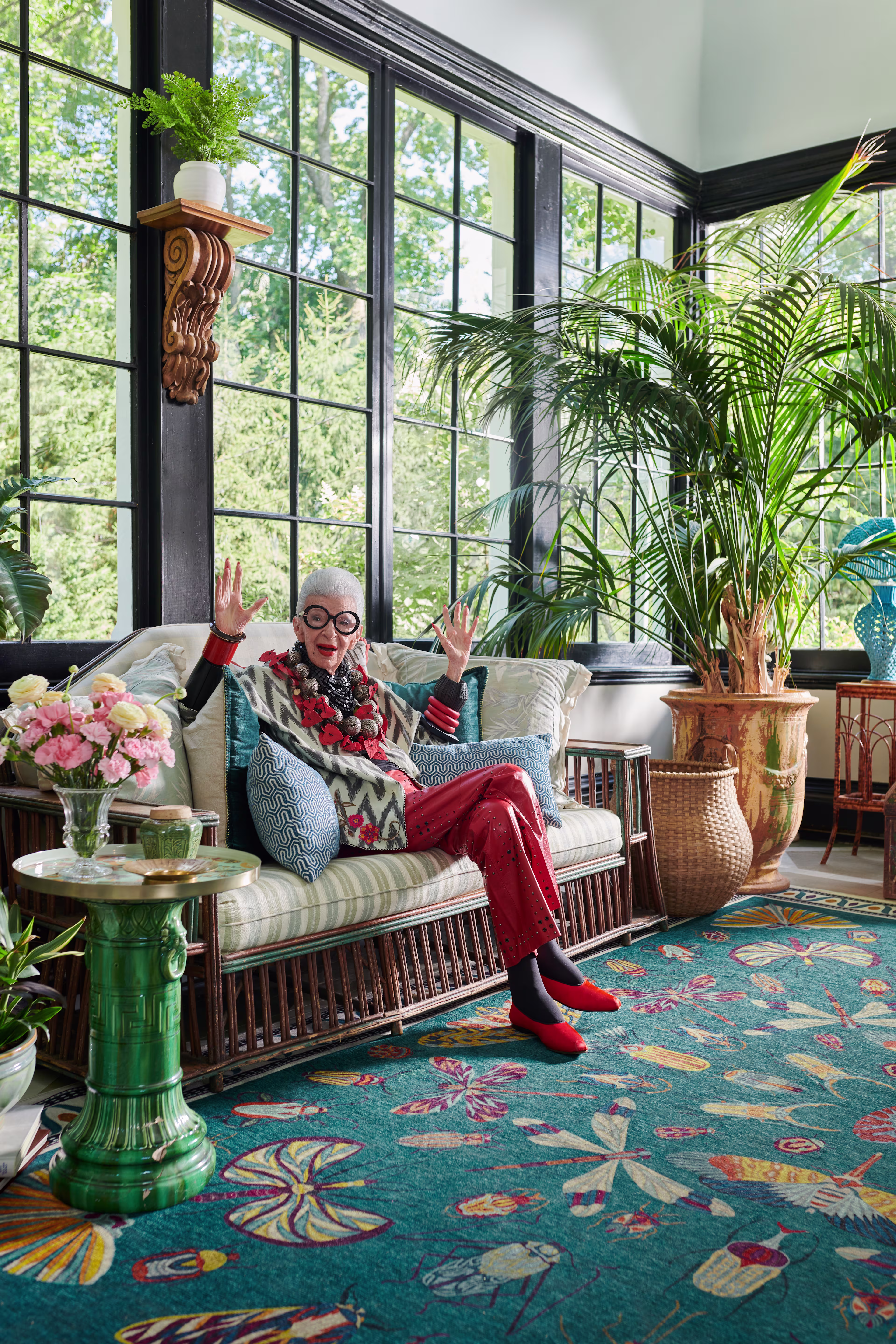 A portrait of Iris Apfel sitting on a striped, wicker-frame sofa in a sunroom filled with plants. She is wearing her signature large round glasses, vibrant red pants, red shoes, and statement necklaces, gesturing with both hands raised. The room features large black-framed windows, a patterned teal rug featuring bird and dragonfly motifs, a green side table with fresh flowers, and a large potted palm.