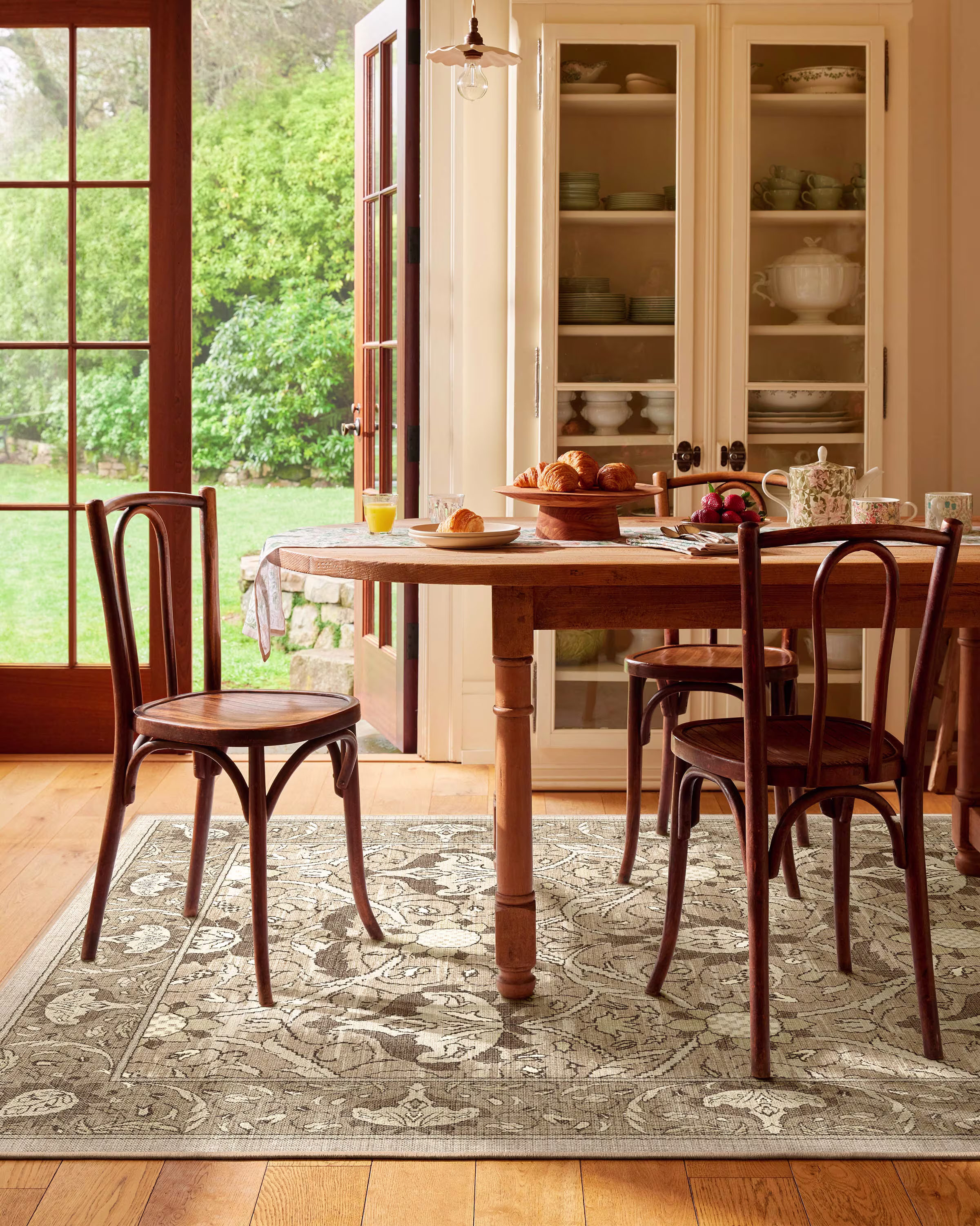 A wooden dining table with three chairs sits on a patterned rug in a sunlit room, with breakfast items and crockery arranged on the table. Glass doors and a china cabinet are in the background.