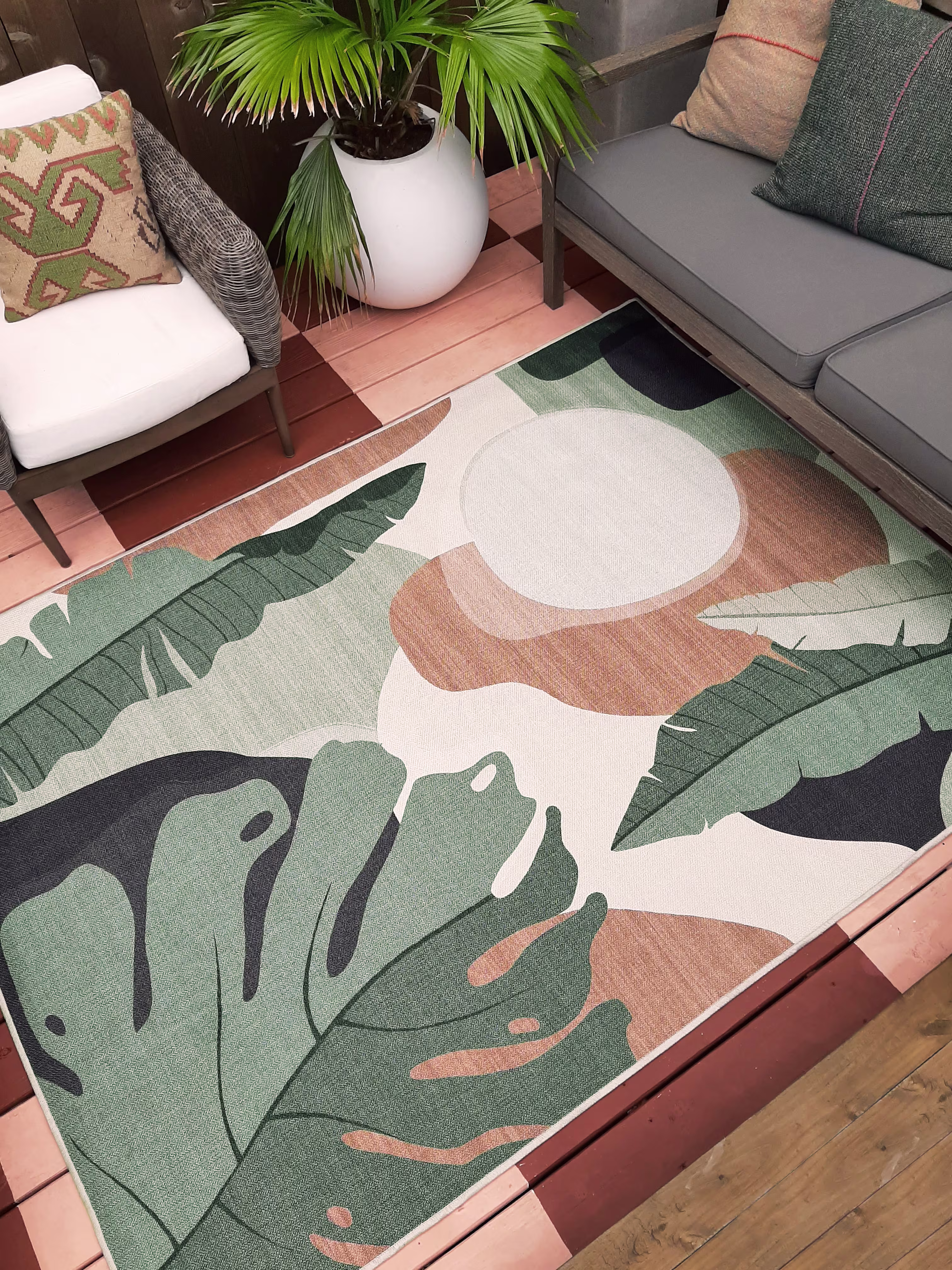 A high-angle shot of the Ruggable Outdoor Havana Desert Sage rug styled on a reddish-brown wood backyard deck. The contemporary rug features large green monstera and palm leaves with abstract tan shapes, placed between grey wicker outdoor lounge chairs with plush white cushions and a concrete accent table.